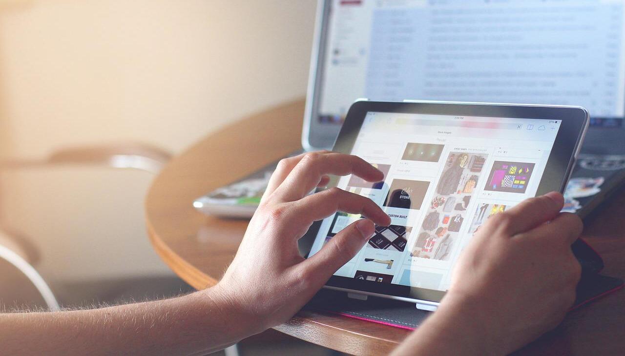 hands scrolling content in a tablet next to a computer screen turned on. 