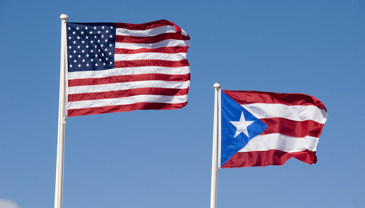 Pictured: U.S flag & P.R flag against the blue sky.