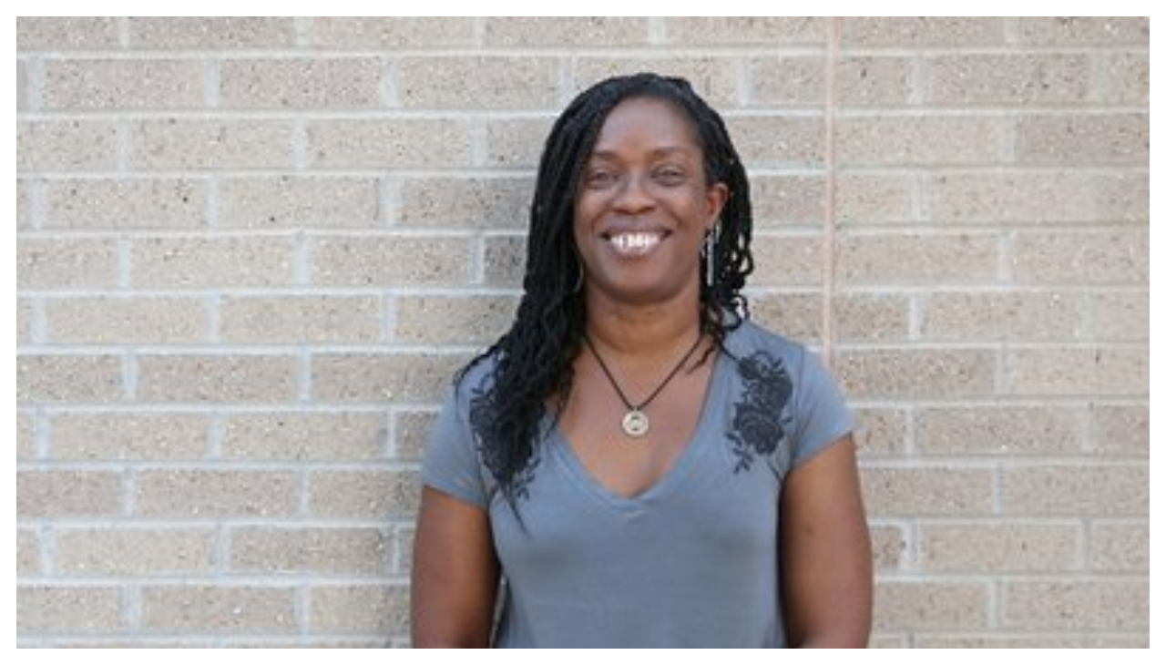 Sheila Rayam standing against a gray brick wall.