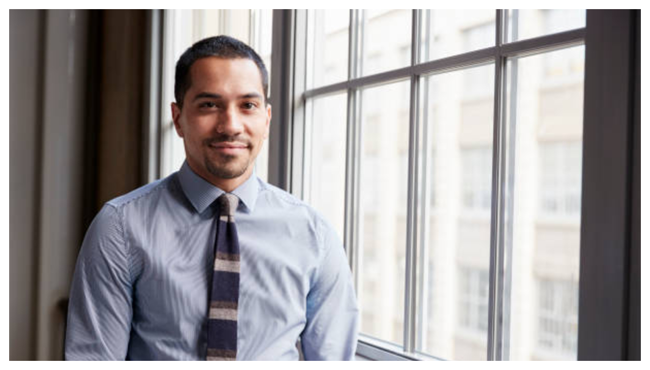 A latino man in business casual dress leaning against a window. He is looking at the viewer.