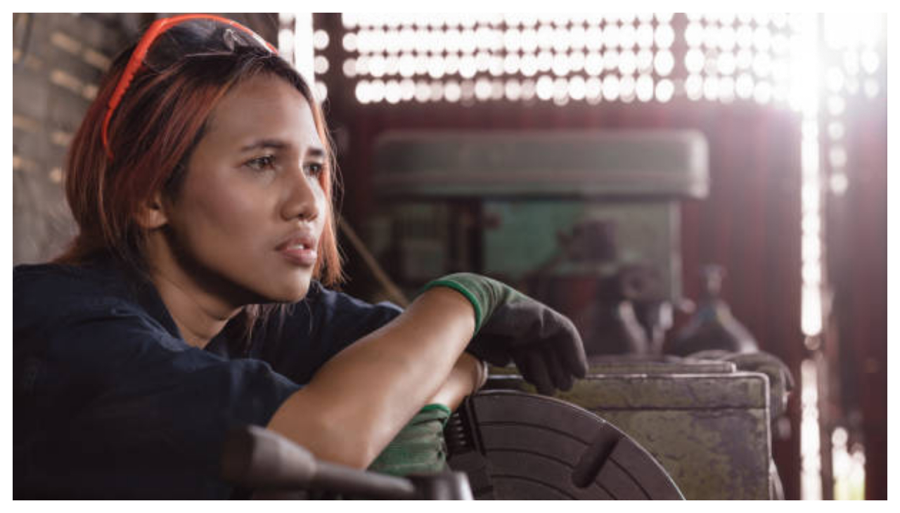 A mexican woman in a warehouse. She is holding a power tool and is sitting down, looking towards the right.