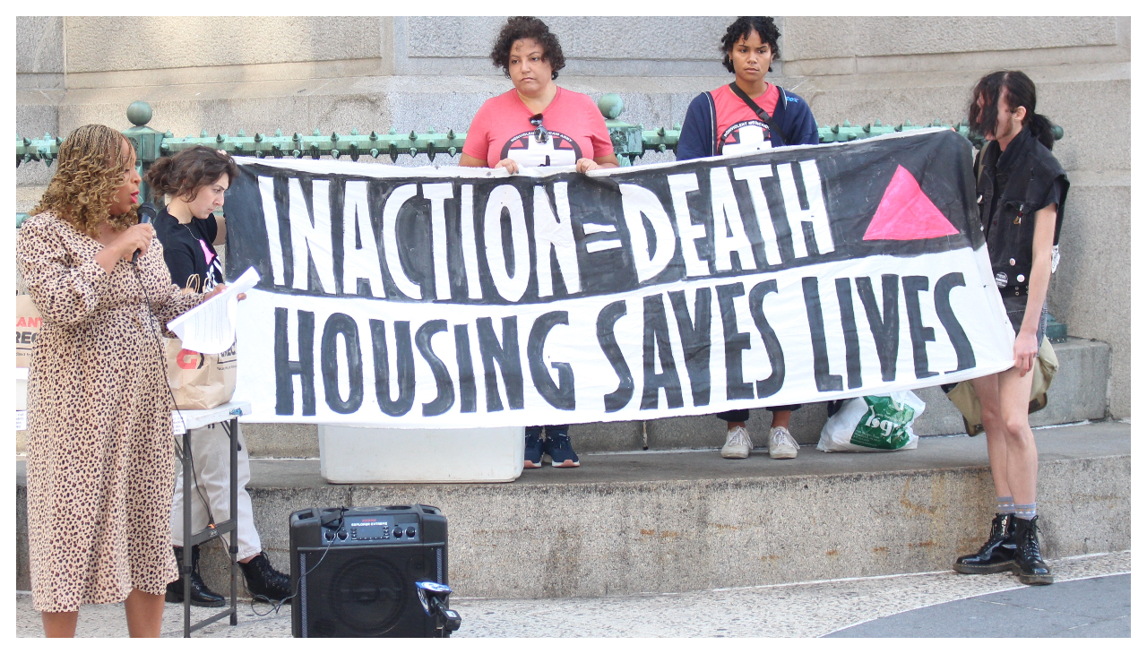 City Councilmember Jamie Gauthier standing in front of a large sign held by four people. The sign reads "Inaction = Death, Housing Saves Lives" with a pink triangle against a black background next to the text.