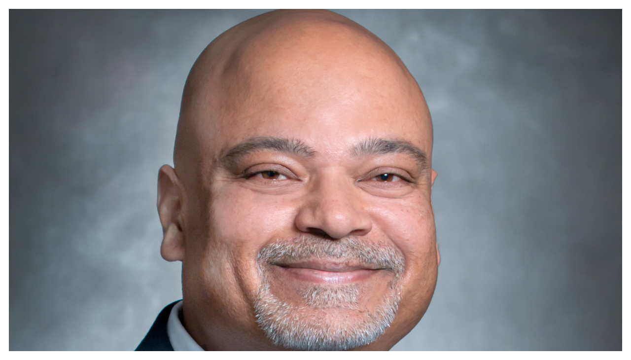 A headshot of Atif Saaed, a bald man with light brown skin. He has a short gray beard and is smiling.