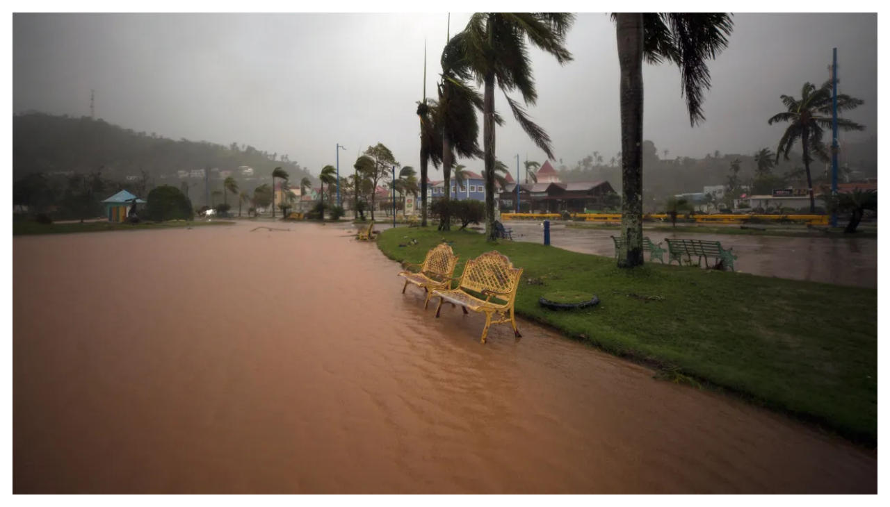 Two yellow benches at the side of a flooded path in Samana, Dominican Republic. The ground is flooded with muddy water. The sky is gray and palm trees are blowing in the wind.