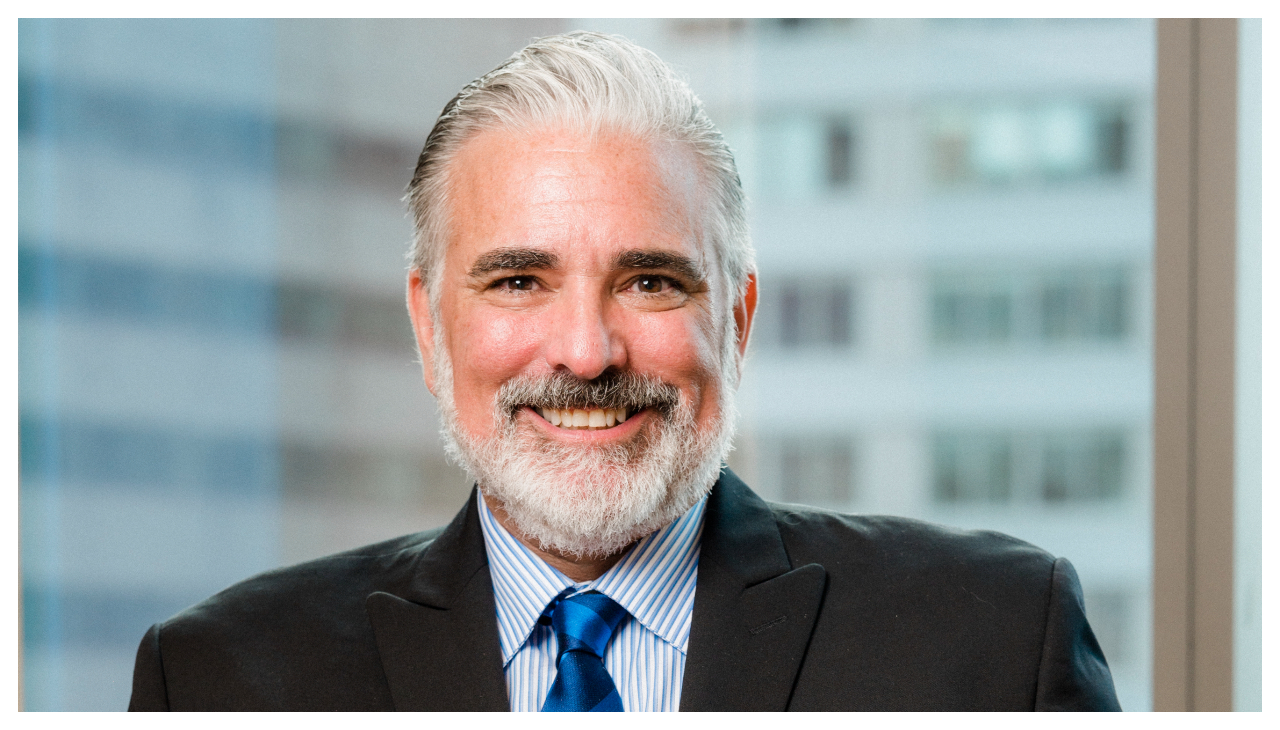 Jose Velez-Silva, a Puerto Rican man with white hair and a beard. He is visible from the shoulders up and is wearing a black suit. He is smiling and looking forward.