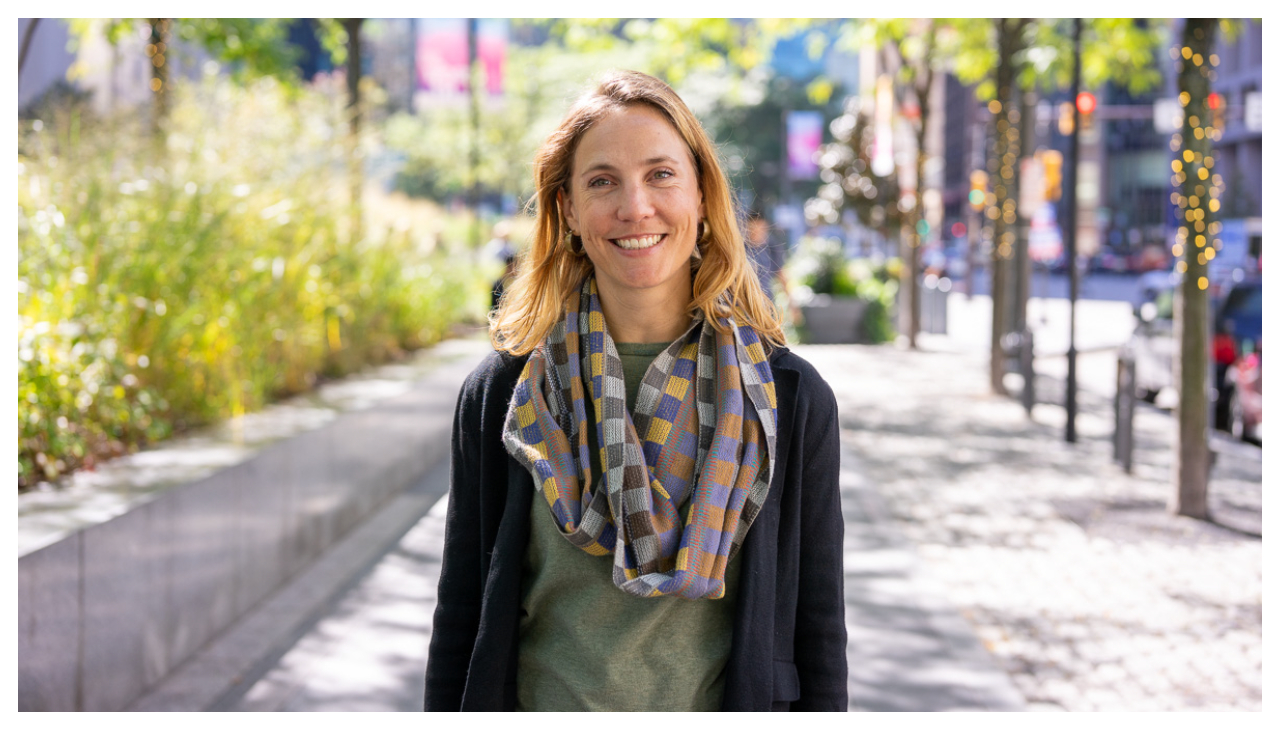 Lily Reynolds, a woman with blonde hair, a green shirt, and a scarf. She is looking at the viewer and smiling.