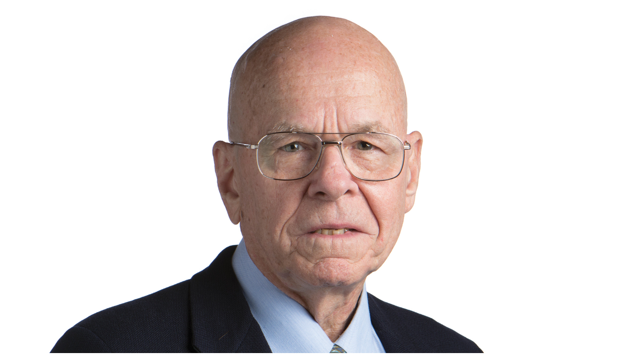 An elderly white man wearing a suit. He is bald and is looking forward.