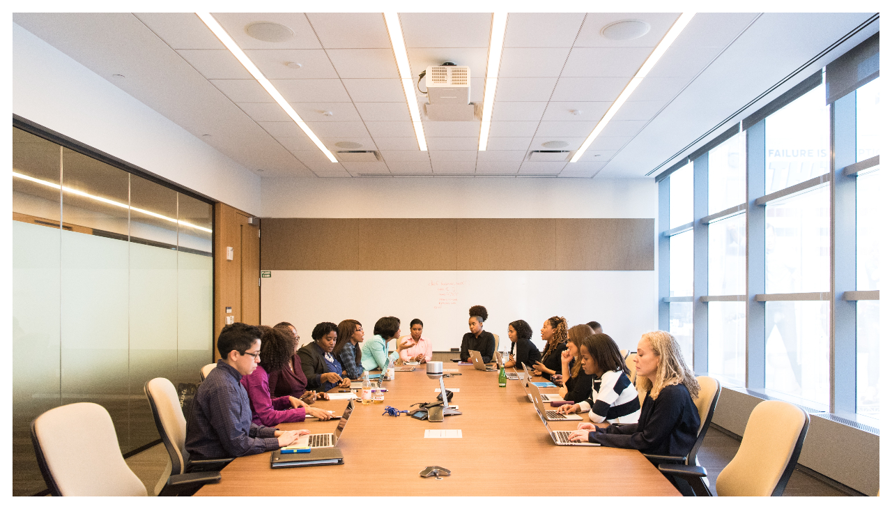 A board room table viewed from one end. At the other end of the table several women are sitting in chairs and looking at a board.