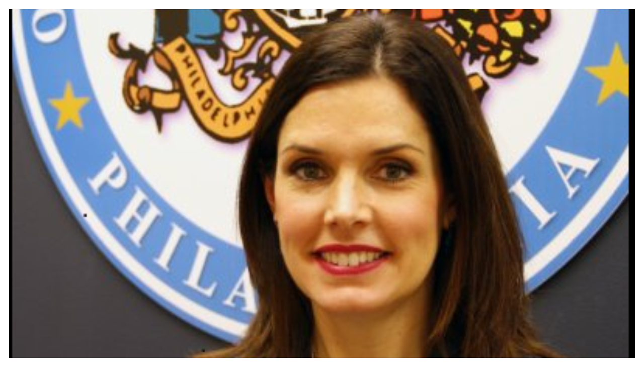 Christy Brady, a white woman with long black hair. She is standing before a seal of the city of Philadelphia. She is looking at the viewer and smiling.