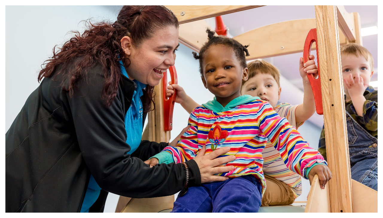 A child prepares to go down a slide as a teacher holds her up.