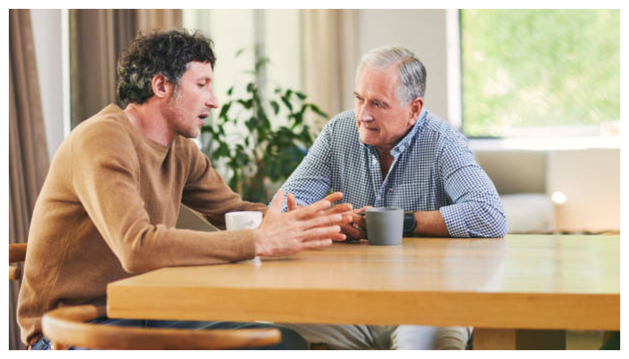 A middle aged white man speaking with an elderly white man at a table. They both look serious and are facing each other.