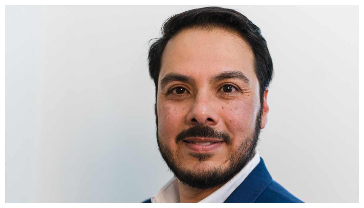 A Latino man with short hair and a beard from the neck up. He is looking at the viewer and smiling.