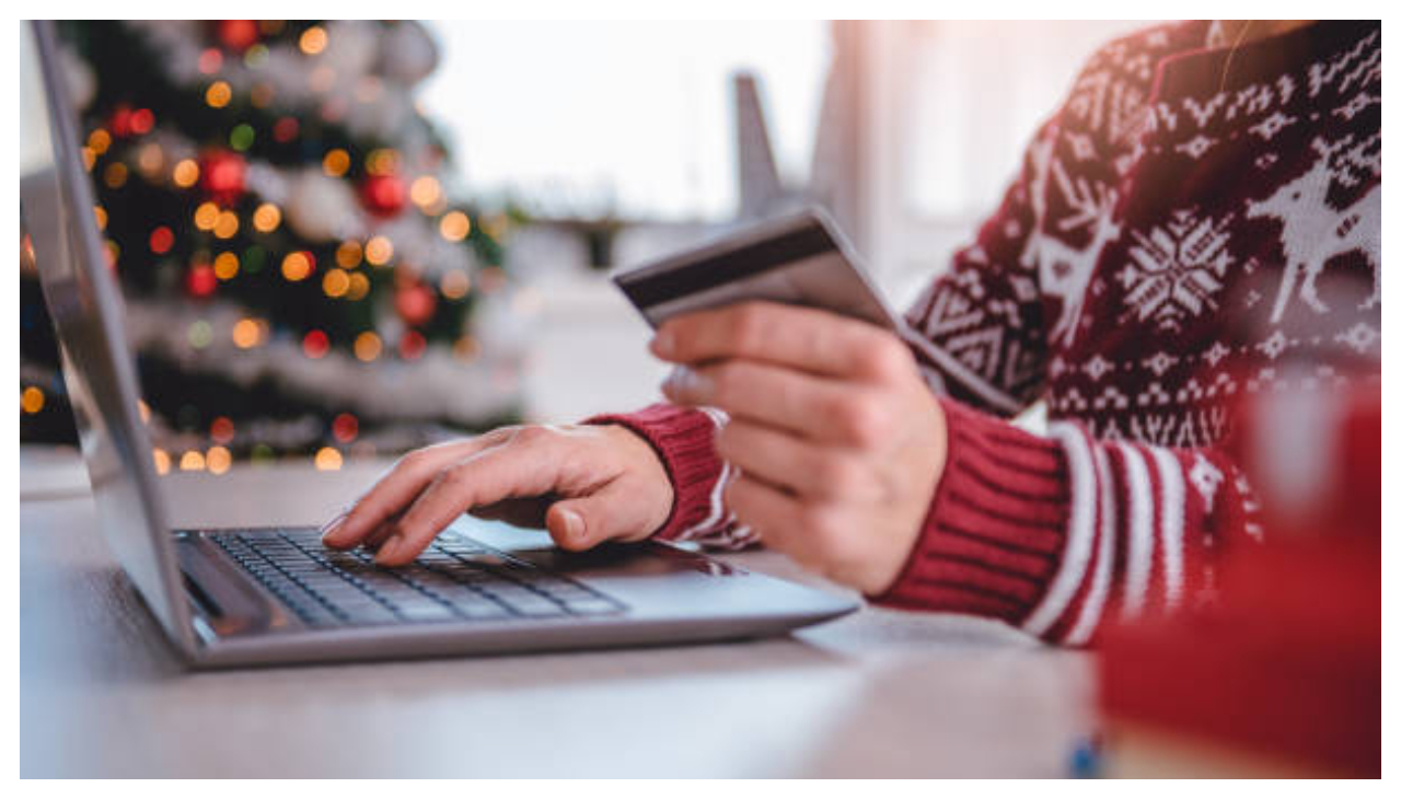 A person in a red Christmas sweater typing at a computer with one hand. In the other they are holding a gift card. A Christmas tree is in the background.