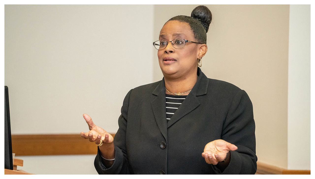 Karla Hill, a woman with light brown skin in a professional jacket and her hair in a bun. She is moving her hands as she speaks at a podium.