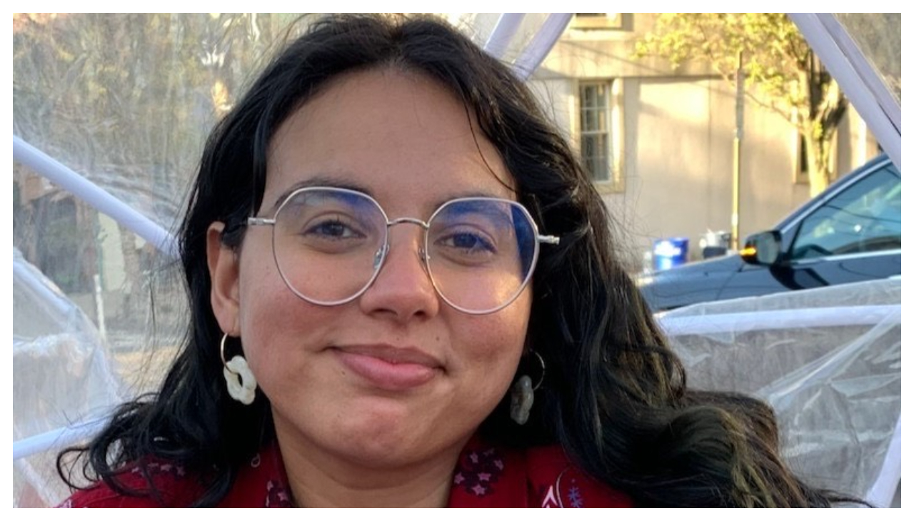 A Zoom call focused on Sabrina Iglesias, a Latina with short black hair, glasses, and a white shirt with a gray pattern. She is smiling.