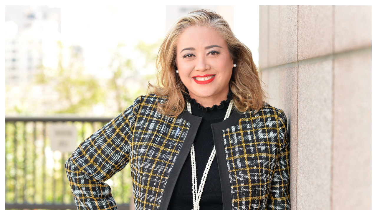 Vanessa Montañez, a Latina women with dark blond hair. She is wearing a yellow and black plaid jacket and is leaning against a wall, facing the viewer.