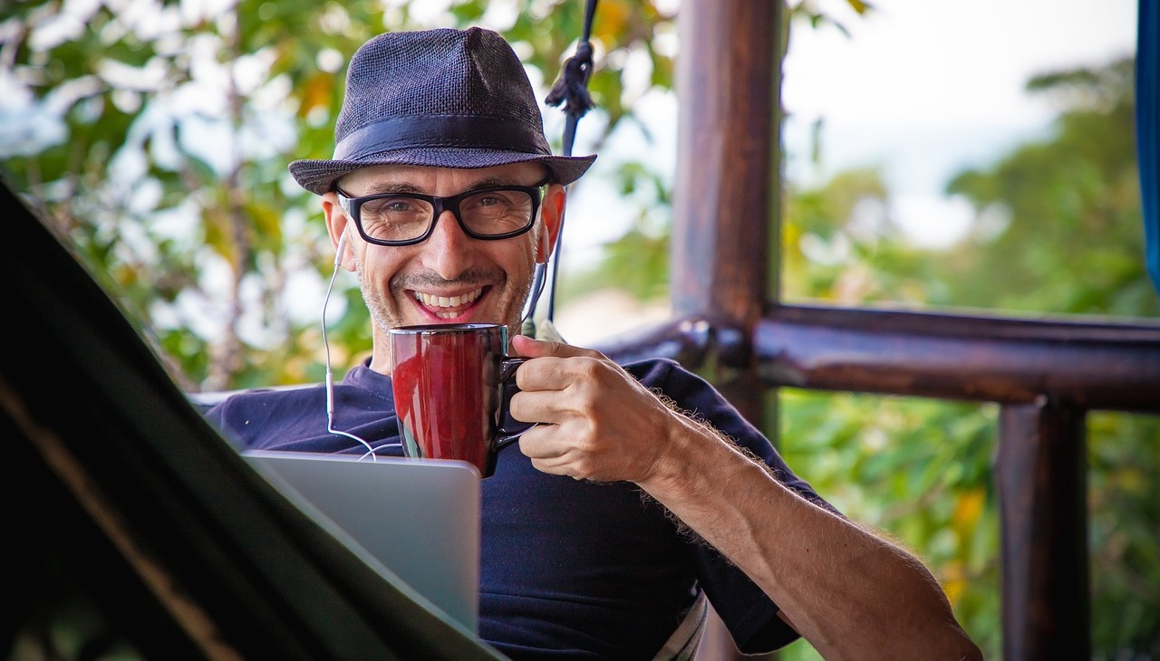 A guy on a hammock working on a laptop and having coffee. 