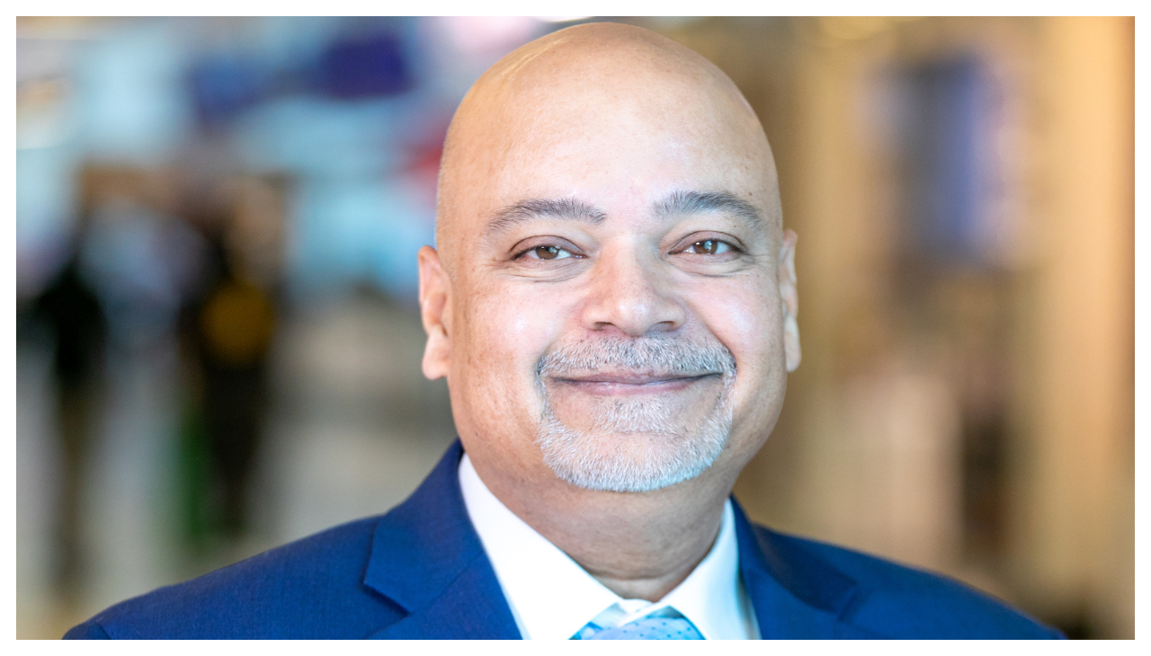 Atif Saeed, a bald Pakistani man with a trimmed and short gray beard. He is shown from the shoulders up wearing a dark blue suit. He is facing the viewer and smiling.