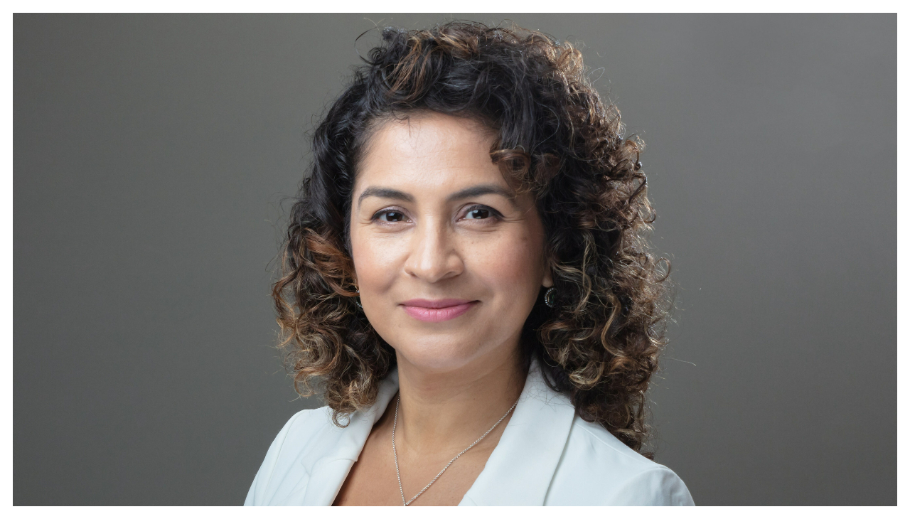 Dr Sarah Lopez from the shoulders up. She is a Latina woman with long curly brown hair. She is wearing a while coat. She is facing the viewer and smiling.