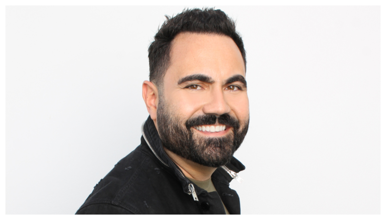 Enriquo Sanchez against a white background. He is a Hispanic man with short black hair and a beard. He is wearing a black jacket and is looking to the viewer from the side.