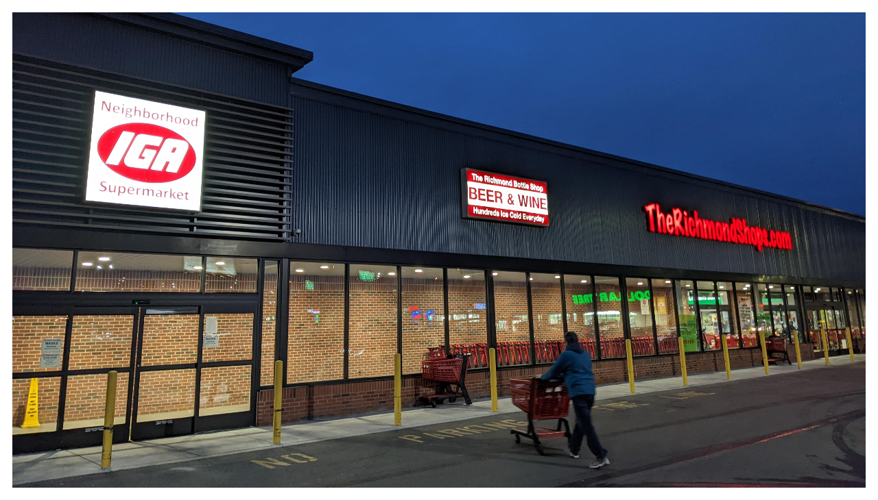 The Richmond Shops grocer at night, lit with a sign saying its name. There is a glowing IGA sign next to it and between the two is a local wine store sign.