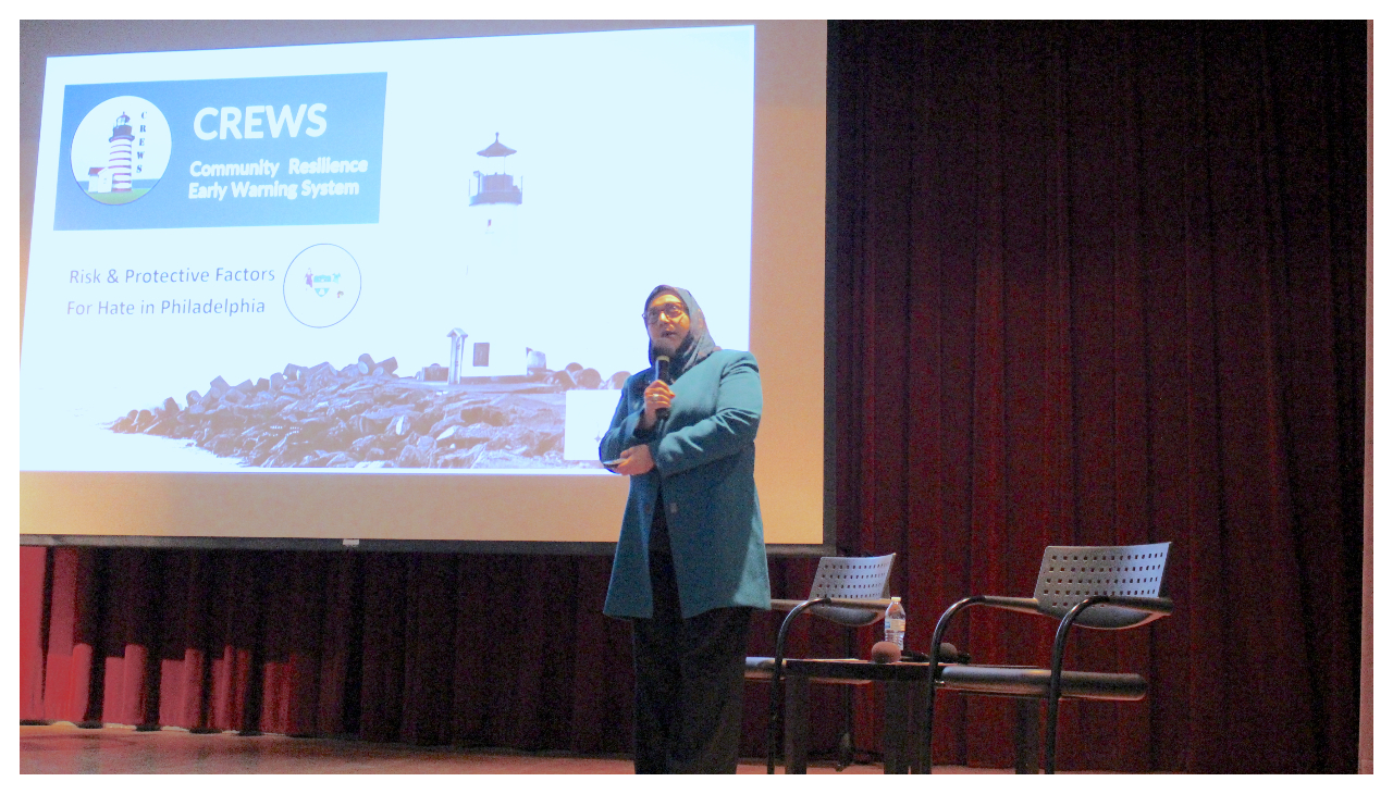 Humera Khan standing on a stage, holding both a microphone and a presentation remote. She is wearing a light blue hijab. Behind her is a projection of a presentation, the slide reading CREWS: Community Resilience Early Warning System. Risk & Protective Factors For Hate in Philadelphia. There is an image of a lighthouse on the slide as well.