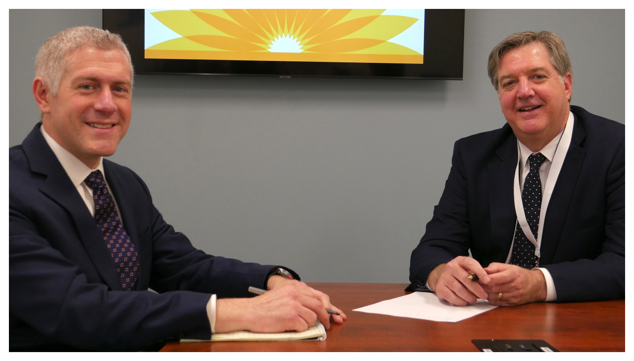 Two men in suits sitting at a table. The first is John Rafferty and the second is Ronan W. Gannon. They are both looking at the viewer and smiling. On the wall behind them a monitor displays the LCH Health's logo.