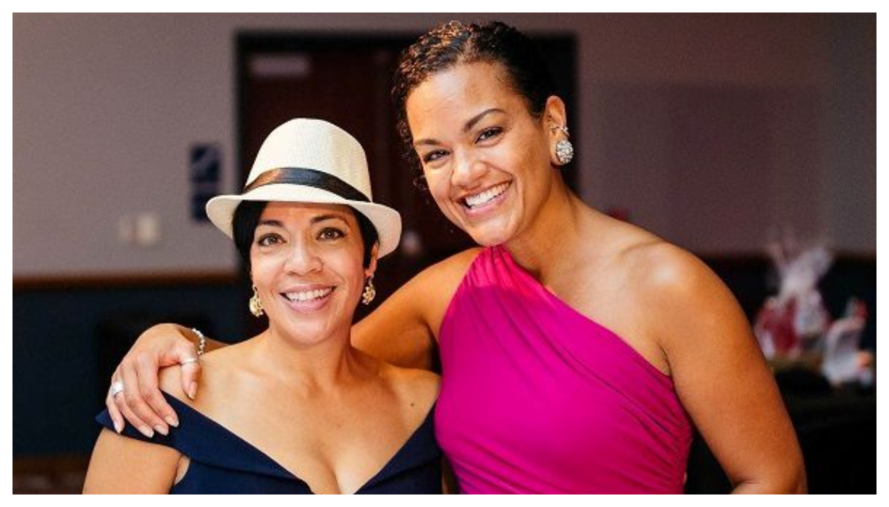 Two women wearing elegant dresses standing next to each other. The woman on the left is wearing a black dress, while the woman on the right wears a pink dress. The woman on the right wrapping her arm around the shoulders of the woman on the left. They are both smiling and looking at the viewer.