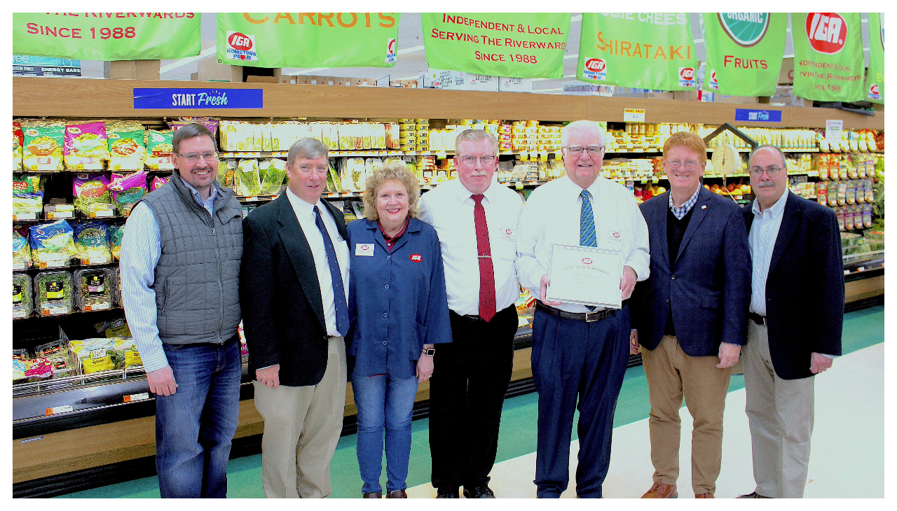 John Hallinan and several employees standing together in the Richmond Shops grocery store. Hallinan is holding an award from the IGA for Retailer of the Year. Behind them are several green banners listing numerous achievements the store has reached.