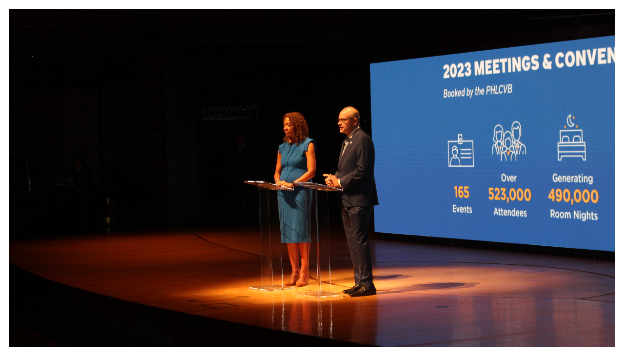 Angela Val and Gregg Caren on stage. Behind them a presentation on the statistics of the tourism industry is displayed.