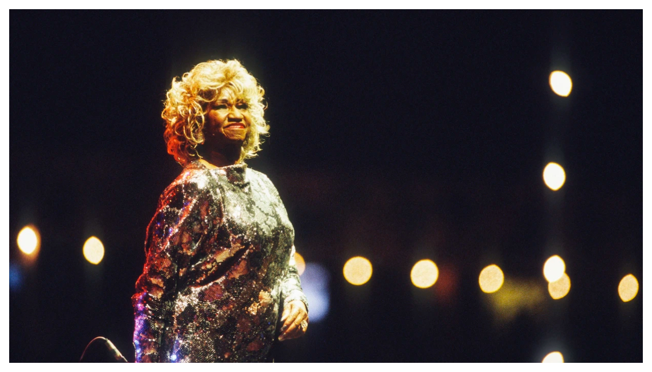 Celia Cruz, an Afro-Latina woman wearing a sparkling gold dress. Her hair is dyed blonde and is looking off towards an audience, holding a microphone at her side. Behind her a dark background is lit by two lines of small, blurry lights intersecting to her right.