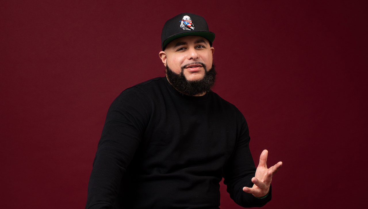 Hector Nuñez standing in the entrance to City Hall subway station . Behind him is a white door. He is wearing a jacket and a baseball cap.