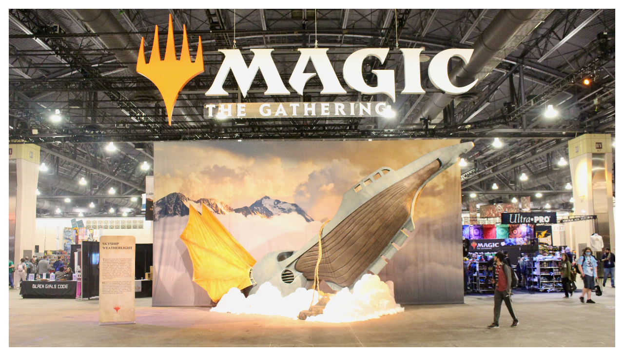 The entrance to the main hall hosting the MagicCon. Hanging from the ceiling is a large sign reading "Magic The Gathering" with the game's logo. On the ground is a large prop depicting a flying ship rising from the crowds. behind it are multiple stalls of card sellers and crowds of people.