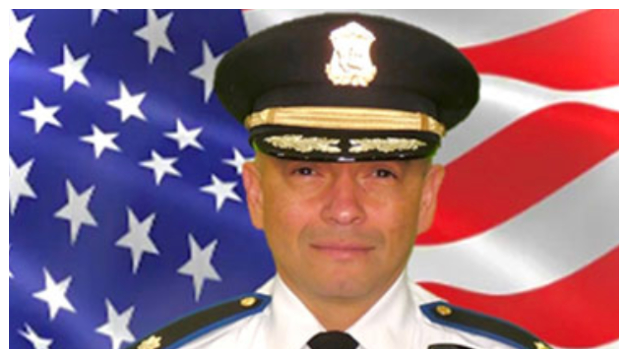 Oscar Perez, a Latino man wearing a white officer's uniform and black cap. Behind him is an American flag.