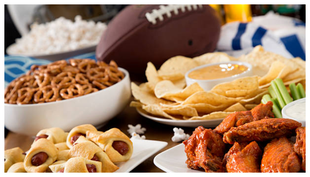 A table laden with food, including chicken wings, hot dog rolls, chips and dip, pretzels, and a football.