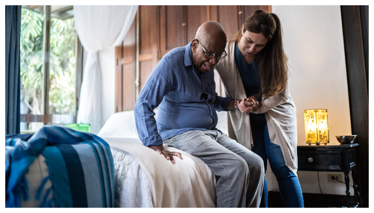 An at home caregiver assists an elderly man in standing as he gets out of bed.