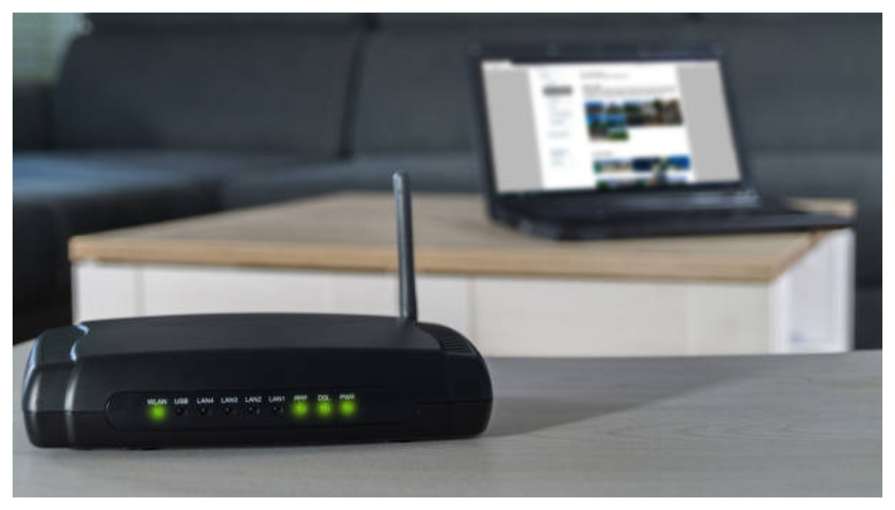 A router sitting on a counter. Behind it is a laptop.