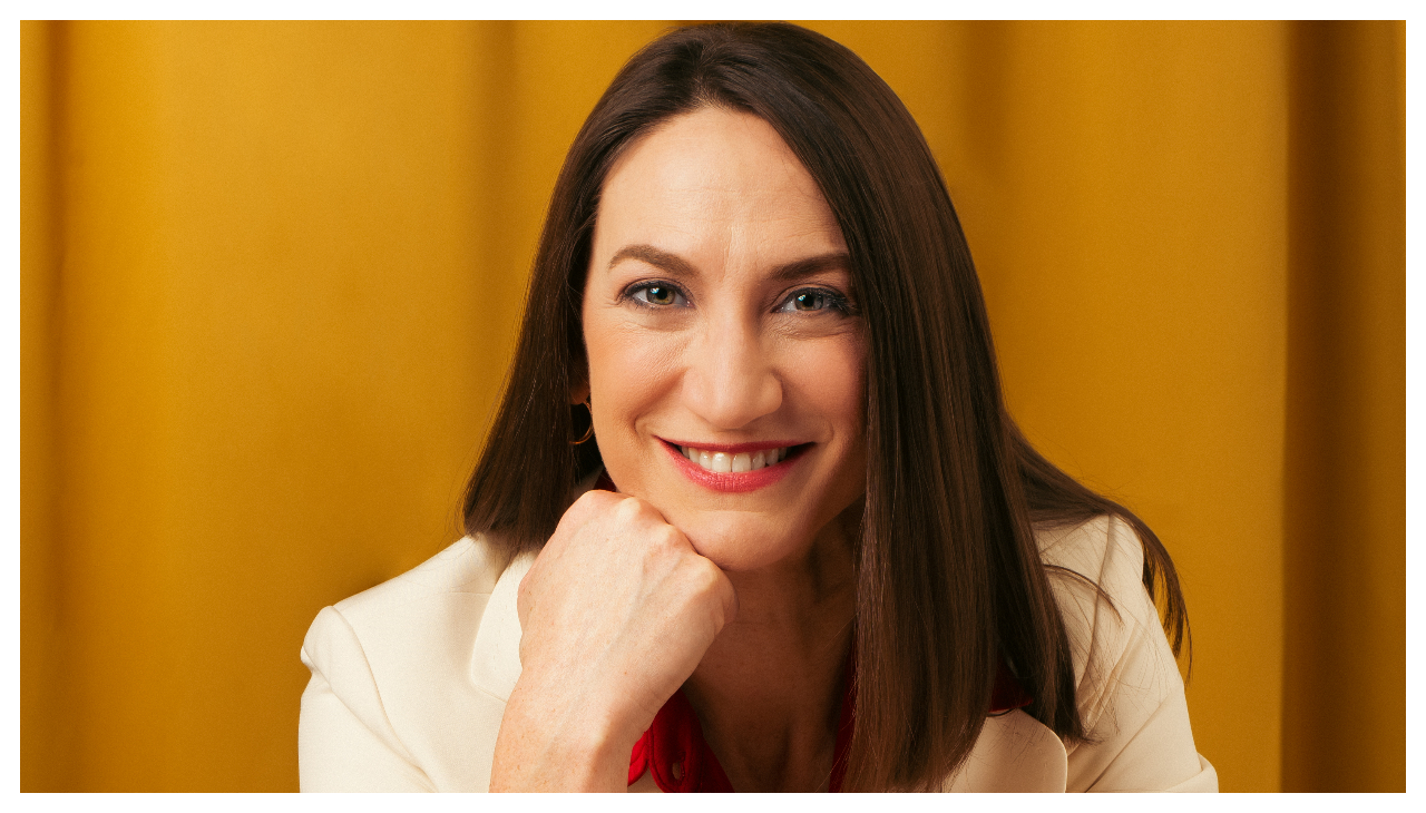 Jenée Chizick-Agüero, una mujer vestida con una chaqueta de abrigo blanca. Ella está apoyada en su mano, mirando al espectador. Ella esta sonriendo.