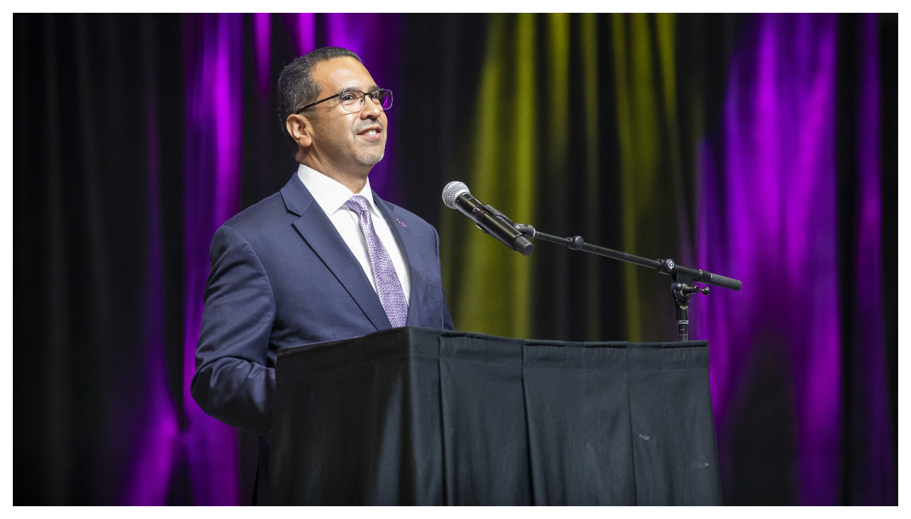 Jose Aviles speaking at a poium. He is wearing a suit and tie and looking to the right towards an audience out of frame. Behind him are purple curtains.