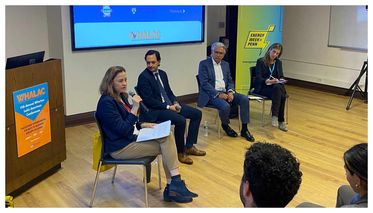 A group of panelists seated before an audience. Beside them is a podium and a projection.