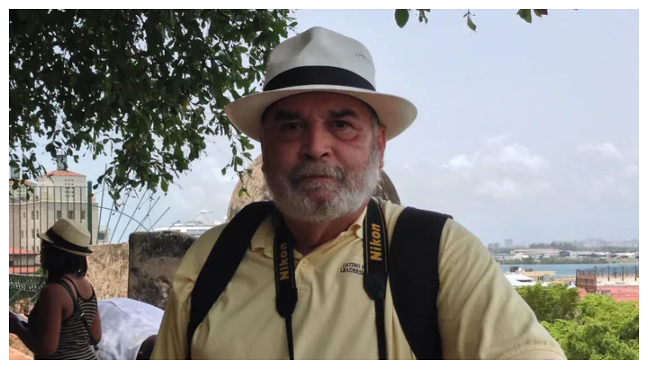 Angel Medina, a Puerto Rican man with short gray hair. He is wearing a tan button up shirt, and a pale hat. He is wearing a backpack and is standing outdoors. He is facing the viewer.