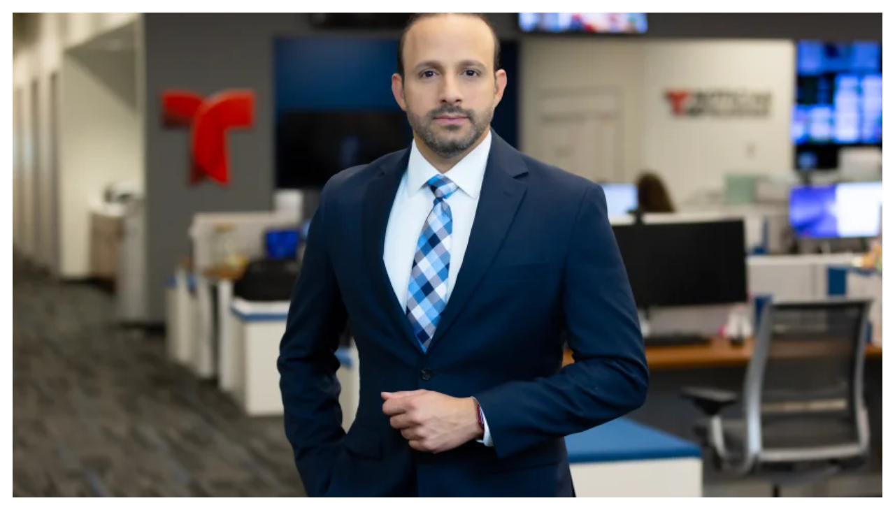 Ariel Rivera-Vázquez, a man wearing a dark suit and a striped tie. He is standing in a newsroom.