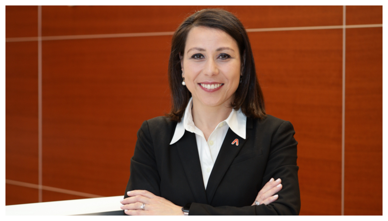 Caroline Vérot Moore, a woman wearing a black women's business suit, standing before a red wall. She has her arms folded in front of her, is facing the viewer, and is smiling.