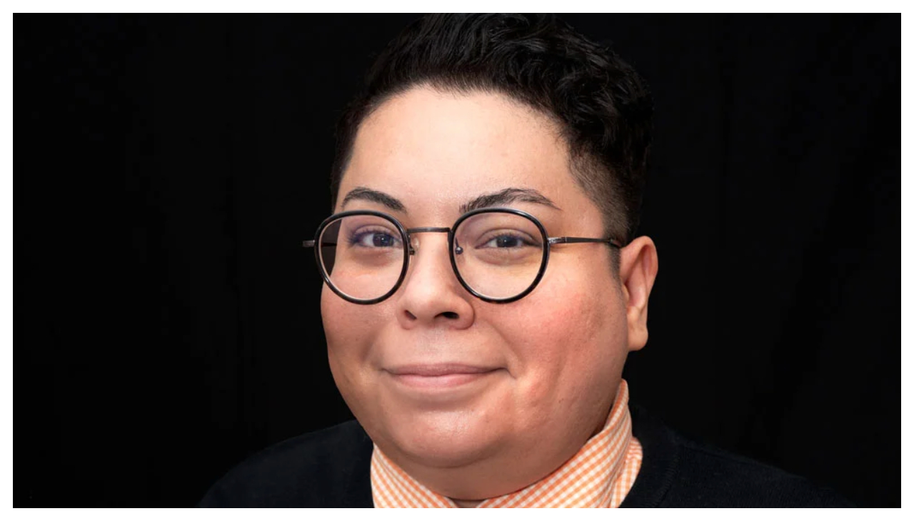 Claudia Zapata, shown from the shoulders up, set against a black background. They are wearing glasses, have dark hair, and are smiling.