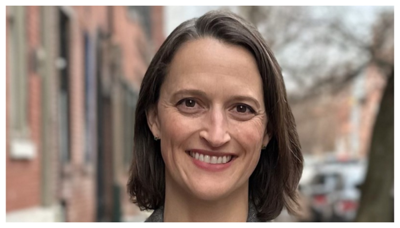 Dr. Megan Todd, from the neck up. A light skinned with neck length straight brown hair. She is facing the viewer and is smiling. Behind her is a line of brick houses.