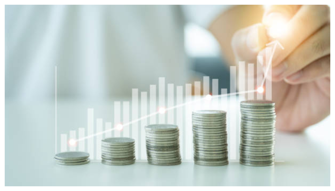 Several stacks of coins next to each other. They start small then increase in size. There is a white bar graph overlaid the coins, representing financial growth. A hand holding a pen traces a line above the bars.