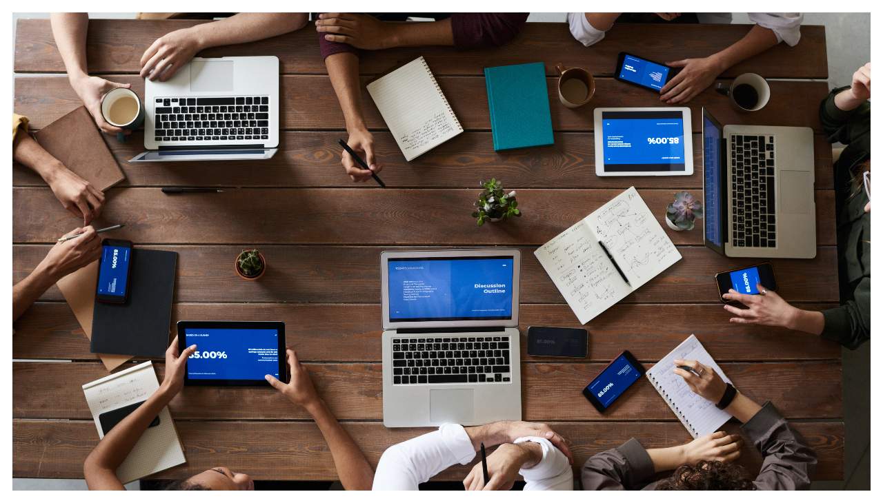 A birds eye view of a group of people seated around a wooden table. Only the people's arms can be seen. There are numerous laptops and notepads in the center. 