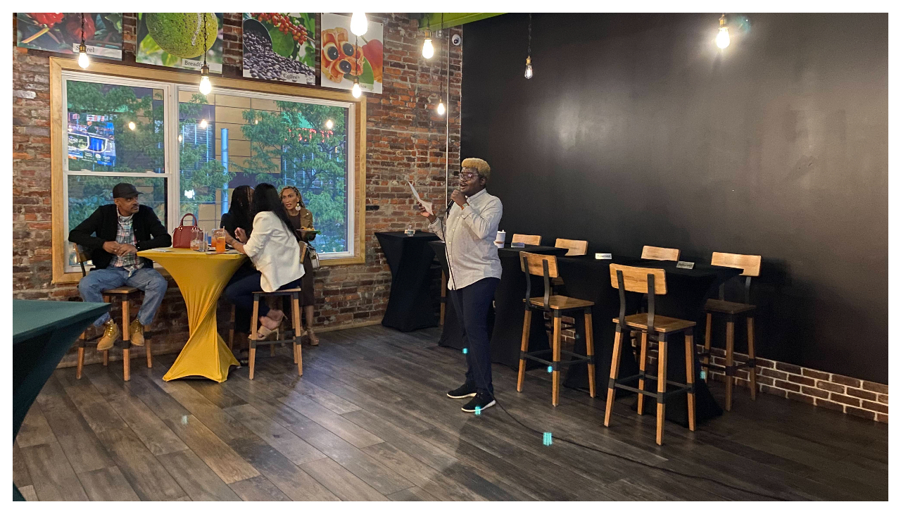 The interior of restaurant bar and grill Irie Entree. Standing at the back of the room before an audience is Chikezie Wood, President of the YCPN, holding a microphone. He is a Caribbean man with dyed blond hair, wearing business casual clothing.