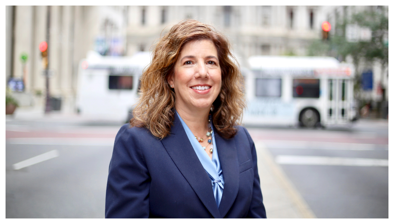 Leslie Richards, a woman with long, light brown hair, shown from the waist up. She is wearing a dark blue blazer with a light blue shirt. Sh is standing in the street with a SEPTA bus behind her, and is facing the viewer, and is smiling.