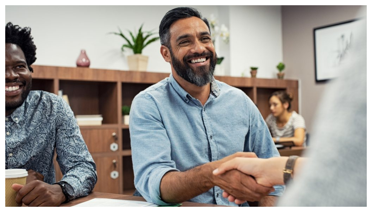 Two men shaking hands, with one out of frame and the other viewed face first. A man to the side sits half in frame. All members are seated in a meeting room and are smiling.